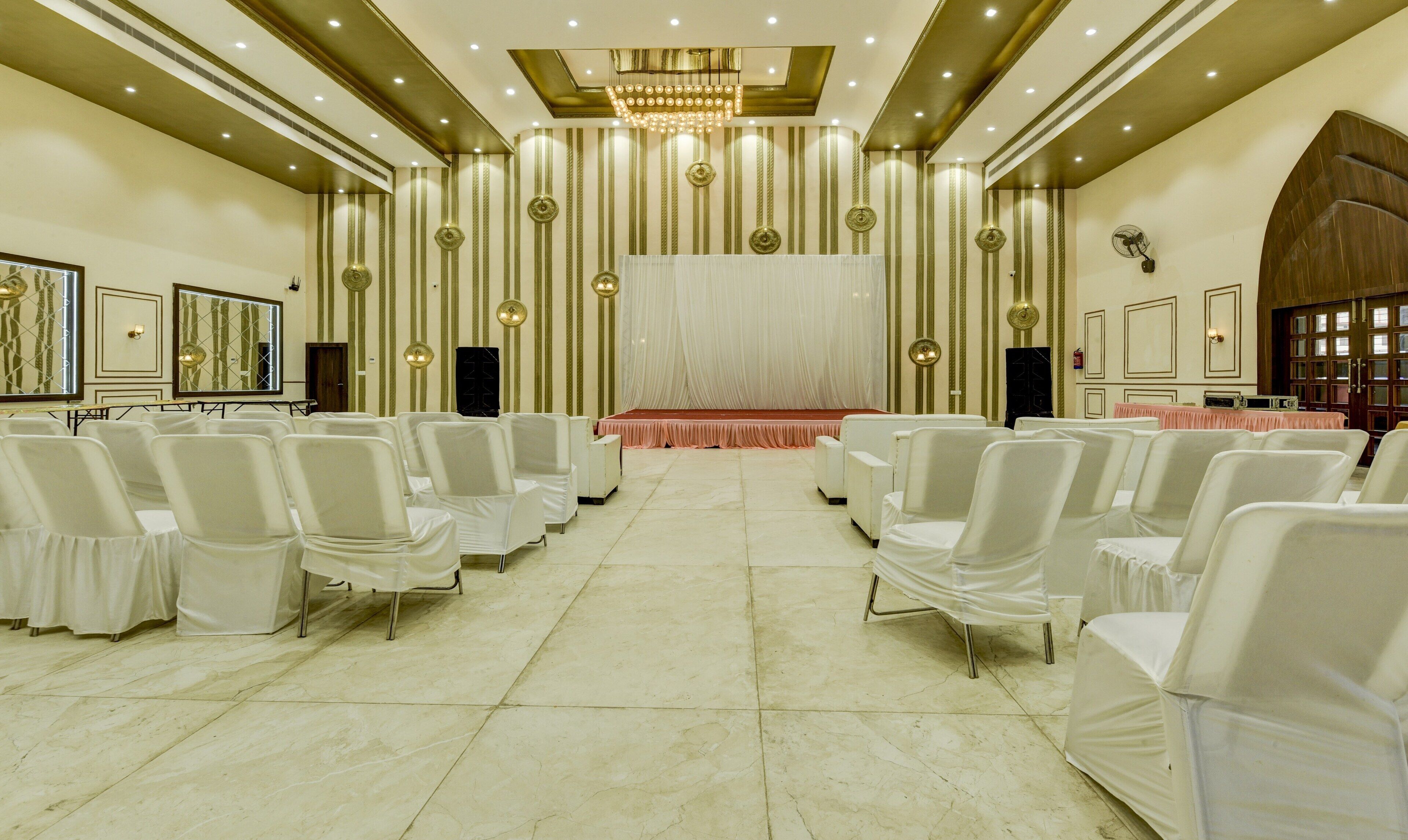 Un salón de conferencias con filas de sillas blancas, un escenario con alfombra roja y una araña de cristal.
