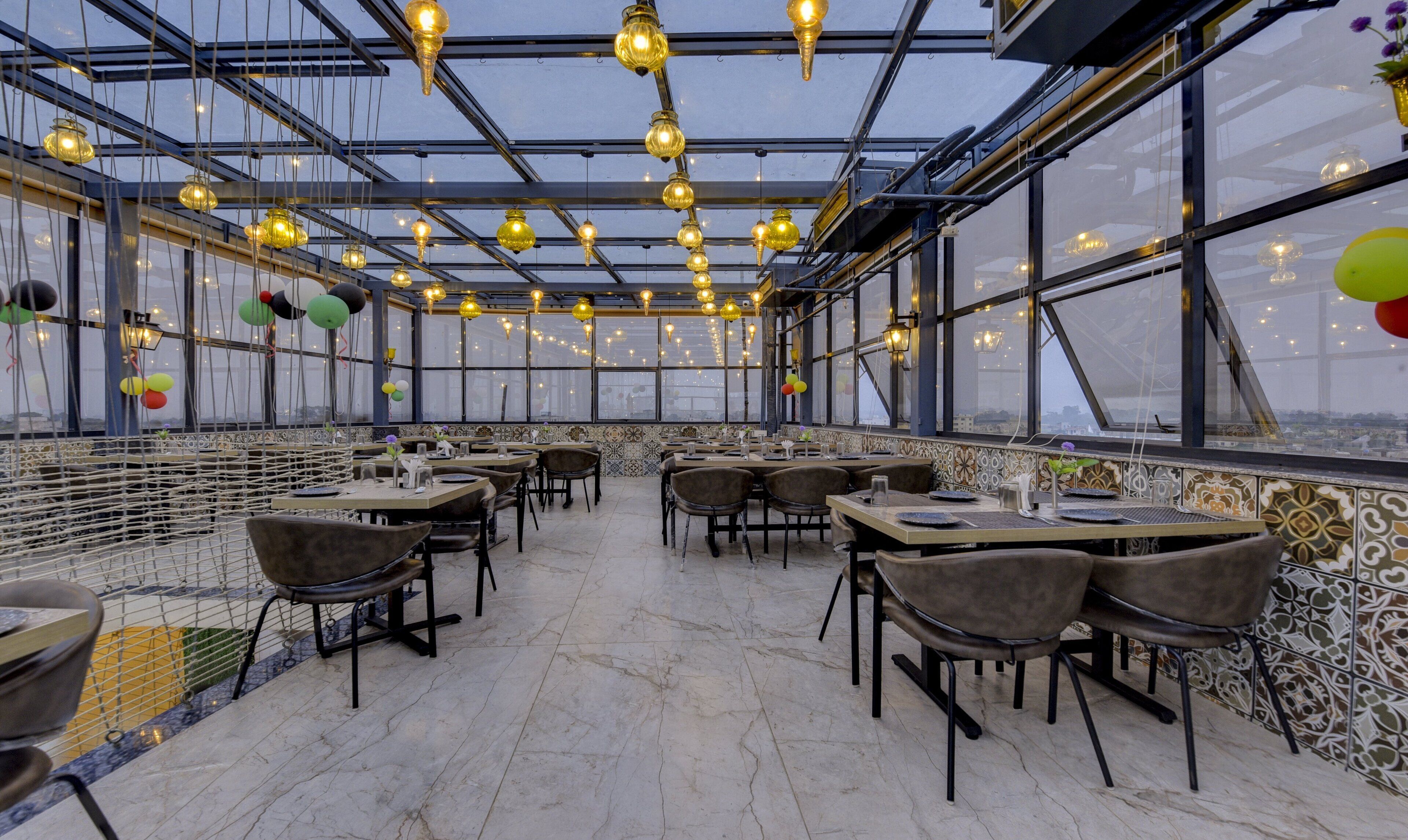 A restaurant with a high ceiling, hanging lights, and a view through large windows. Tables and chairs are arranged for dining.
