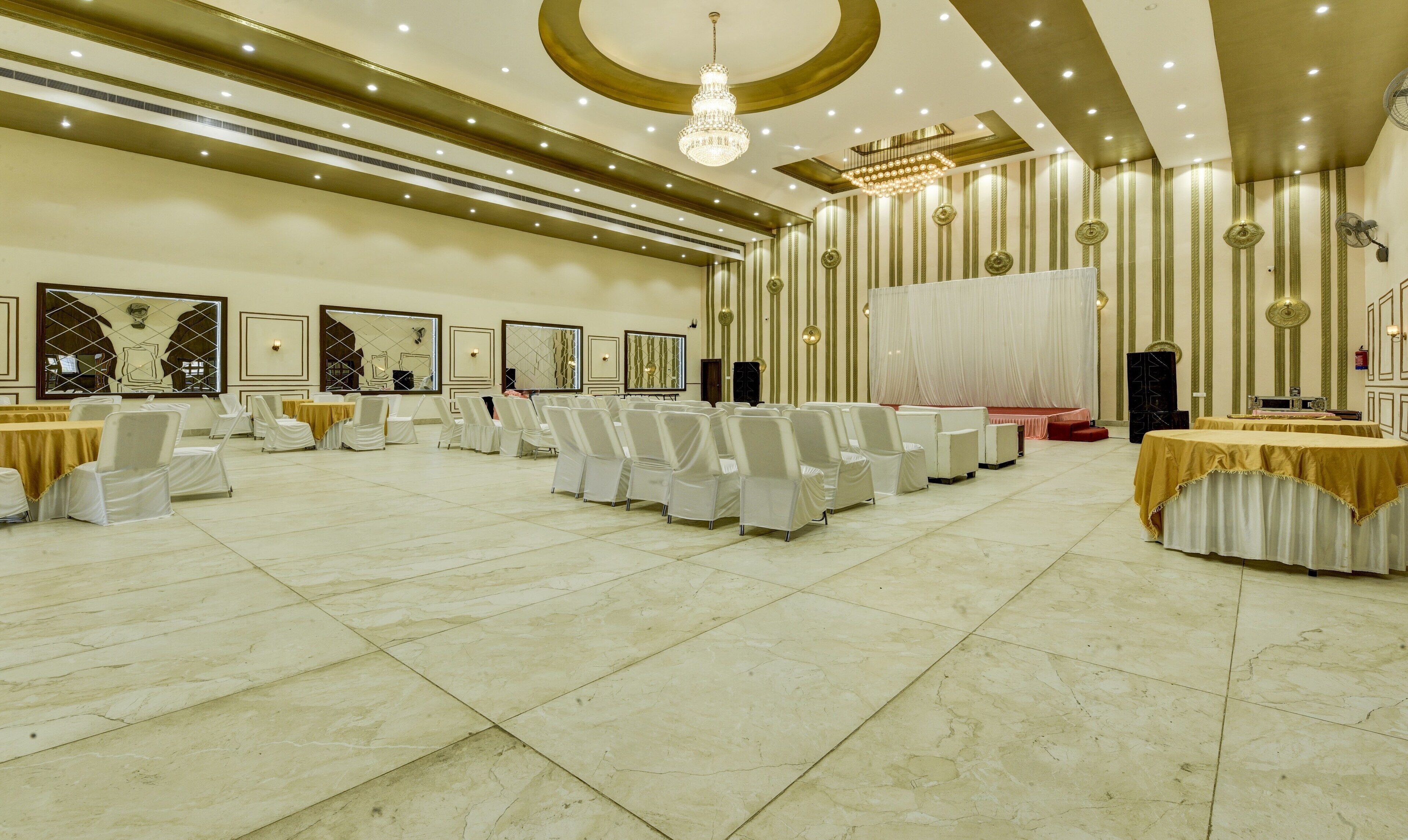 A spacious banquet hall with rows of white chairs, a stage with a red backdrop, and a chandelier.