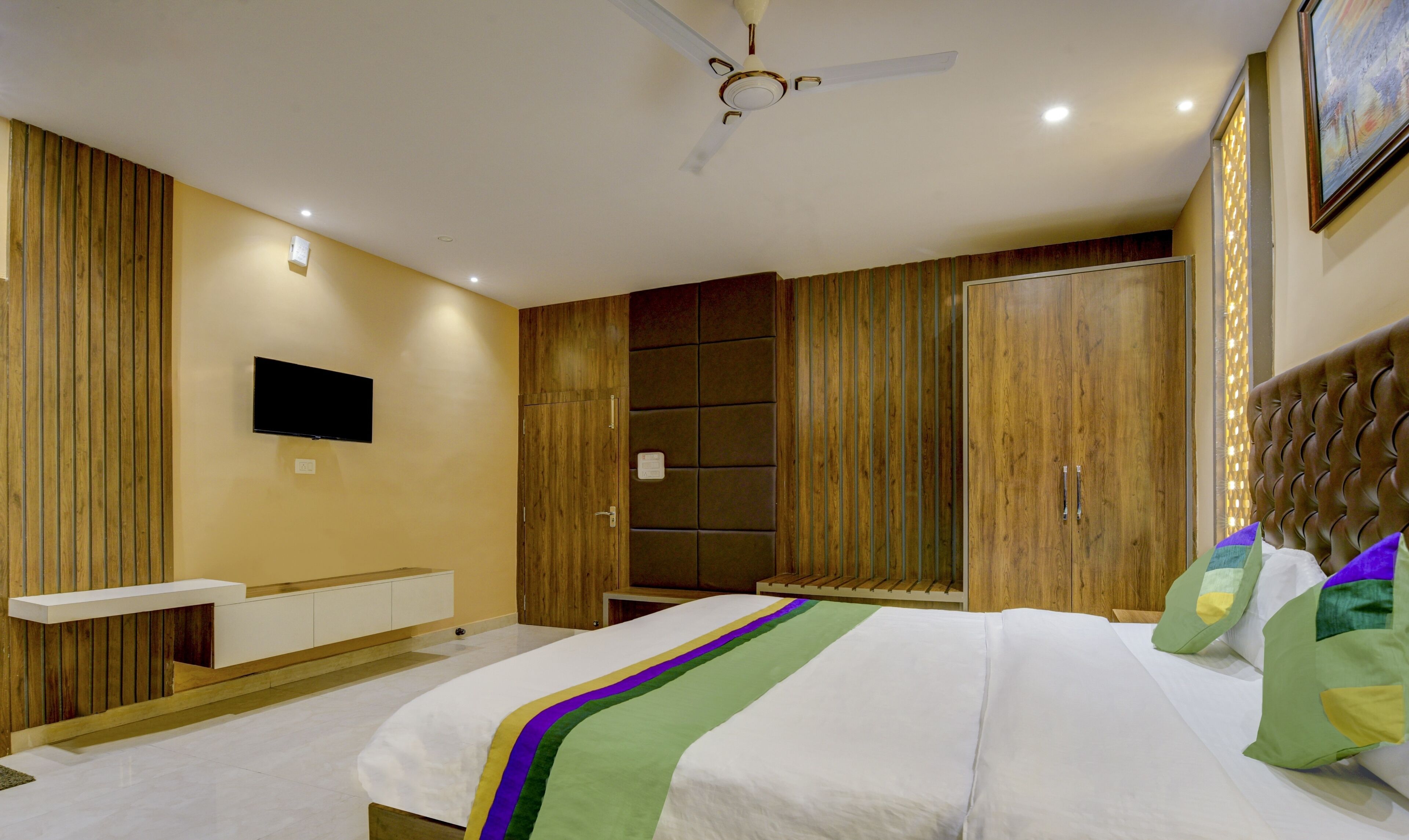 A modern bedroom with a bed, a flat-screen TV, wooden paneling, a ceiling fan, and a large wardrobe.