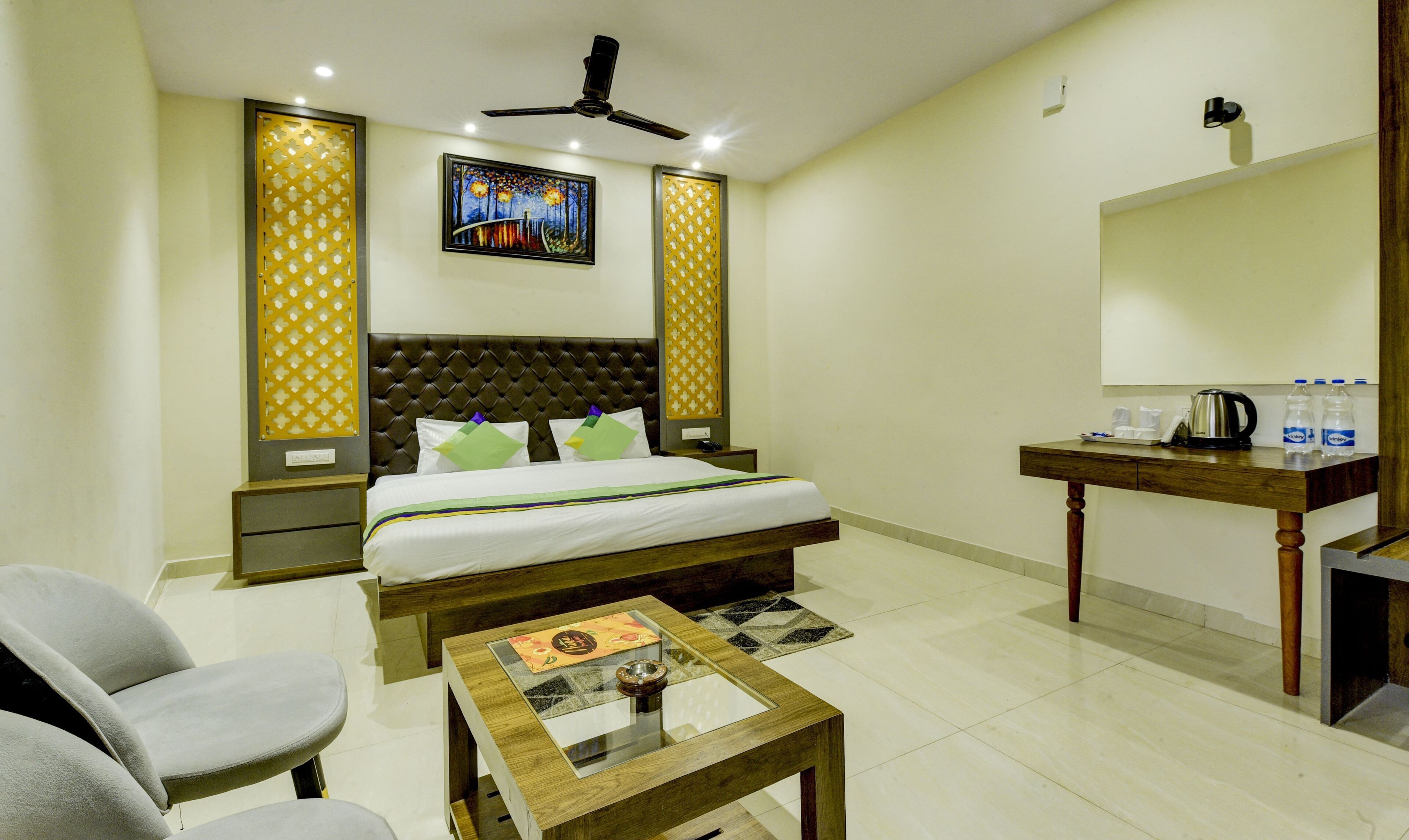 A hotel room with a bed, a desk, a chair, a ceiling fan, and a painting on the wall.