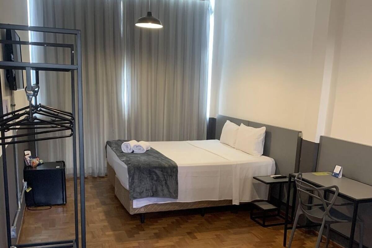 A hotel room with a bed, a desk, a chair, a hanging light fixture, and a window with curtains.