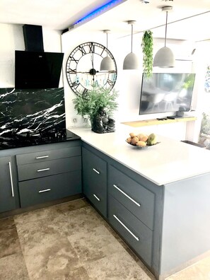 A modern kitchen with a black stove, grey cabinets, a large clock, and a TV on the wall. - Villa vue mer (Collectivité de Saint-Martin)