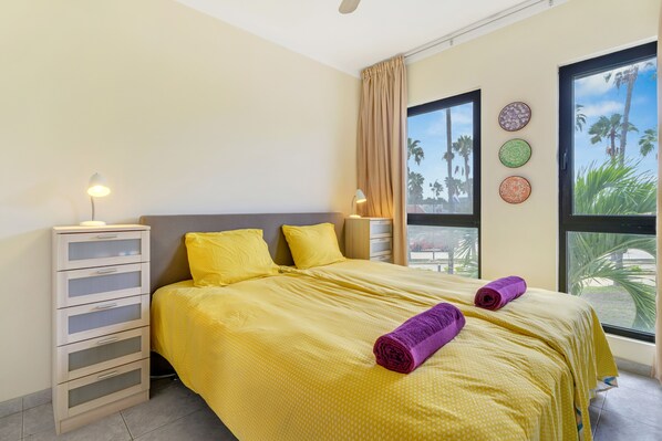 A hotel room with a bed, a nightstand, a lamp, and a window with a view of palm trees. - Caribbean Court - Harbor View Apartment (Kralendijk)