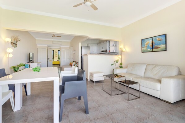 A modern living room with a white sofa, a dining table with chairs, a coffee table, and a ceiling fan. There is a kitchen area with cabinets and a countertop visible in the background. - Caribbean Court - Harbor View Apartment (Kralendijk)
