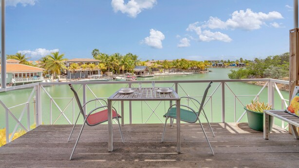 A wooden deck with a table and chairs overlooking a body of water, surrounded by palm trees and buildings. - Caribbean Court - Harbor View Apartment (Kralendijk)