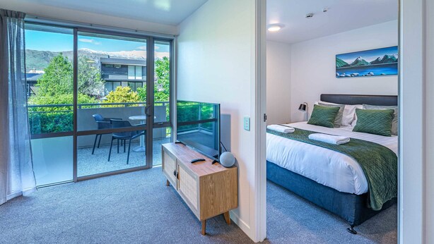 Ett modernt hotellrum med en stor skjutdörr i glas som leder ut till en balkong, en säng med grönt sänglinne och ett litet nattduksbord med en lampa.