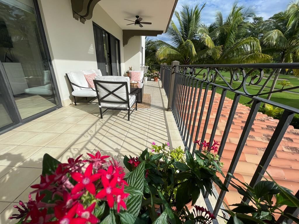 Ein Balkon mit einer weißen Couch und einem Tisch, einem Metallgeländer und Blick auf einen Garten mit Palmen.