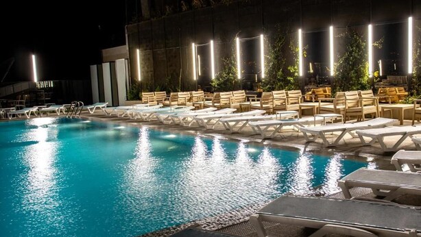 A nighttime pool area with lounge chairs and tables, surrounded by a building with vertical light fixtures. - Hotel Beaumonde Garden (Tbilisi)