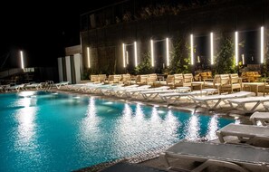 Ein Poolbereich bei Nacht mit Liegestühlen und Tischen, umgeben von einem Gebäude mit vertikalen Leuchten.