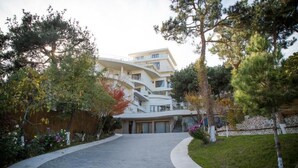 A modern building with a curved facade, a central staircase, and a paved entrance area with trees and shrubs. - Hotel Beaumonde Garden (Tbilisi)