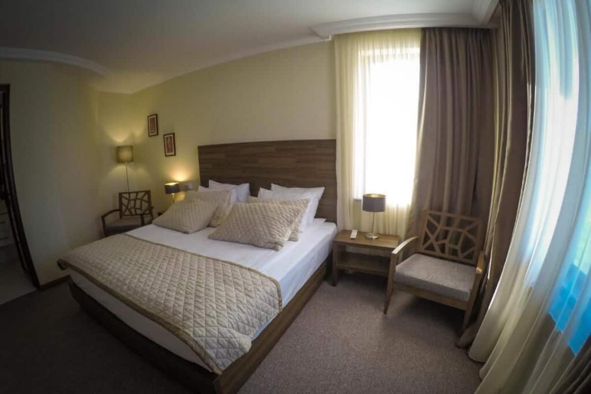 Ein Hotelzimmer mit einem großen Bett, einem hölzernen Kopfteil, einem Nachttisch mit Lampe, einem Sessel und einem Fenster mit Vorhängen.