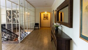 A hallway with a wooden floor, a staircase with glass railings, a black chair, a wooden console table with a vase, and a framed painting on the wall. - Hotel Beaumonde Garden (Tbilisi)