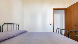 A single bed with a grey mattress and metal headboard in a room with white walls and a wooden door. - 2141 Villetta Roberta - Piano Terra by Barbarhouse (Nardò)