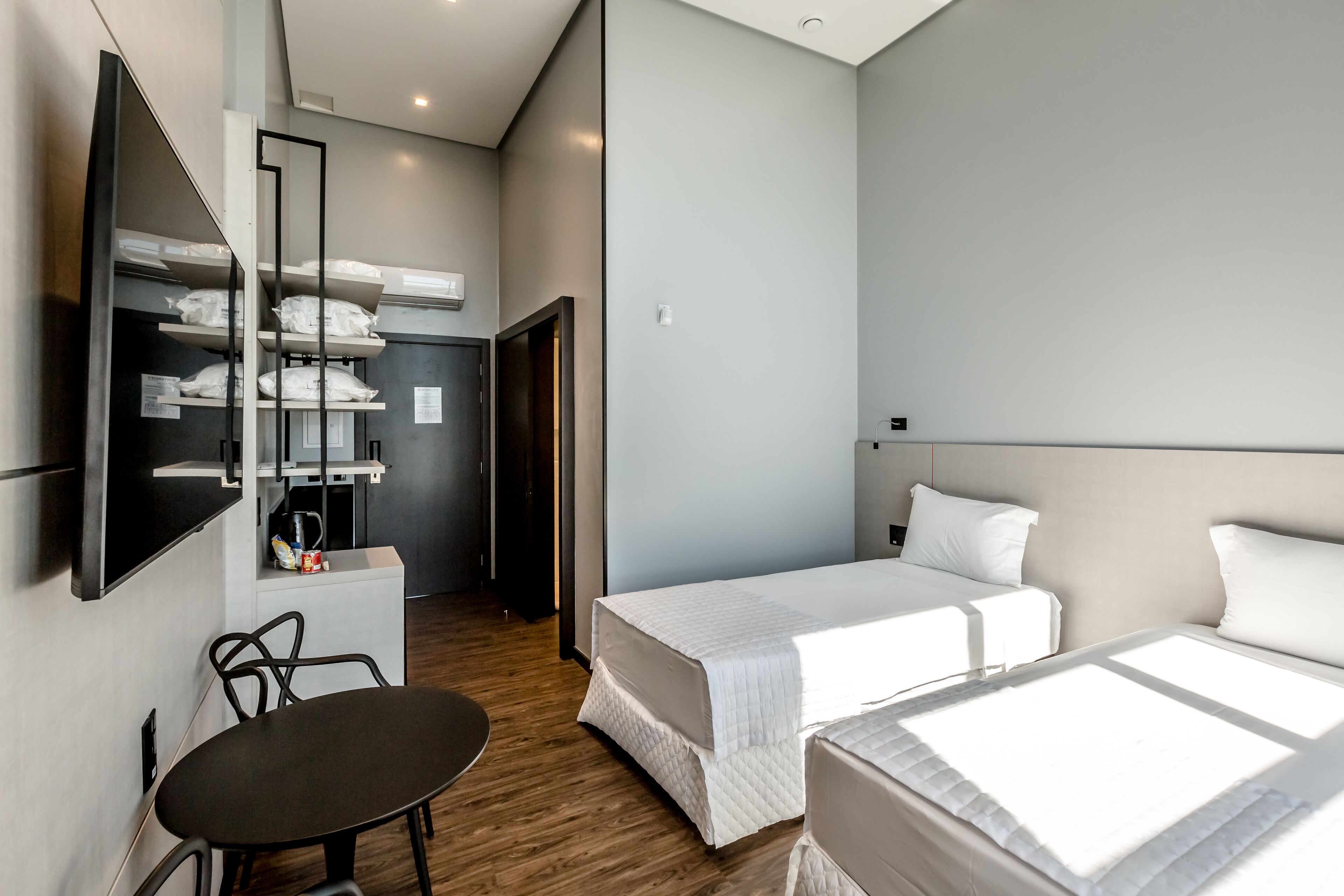 A hotel room with two beds, a TV mounted on the wall, a small table, and a kitchenette area with shelves.