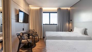 Habitación de hotel con dos camas, televisión montada en la pared, una mesa de comedor con sillas y una ventana con cortinas.
