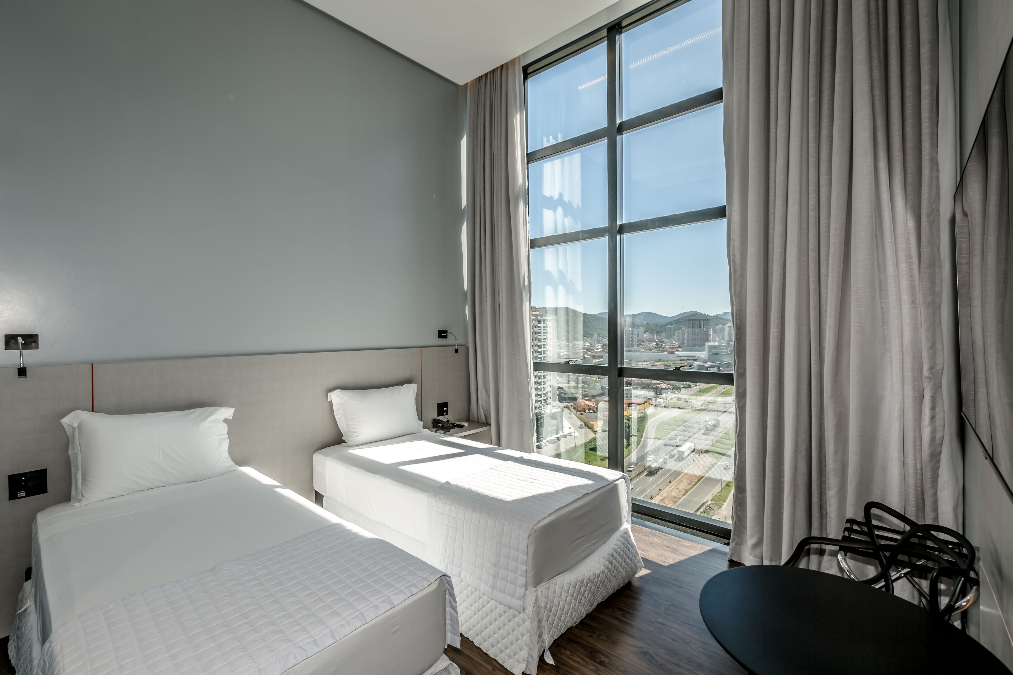A modern hotel room with two beds, a large window with a city view, a desk, and a chair.