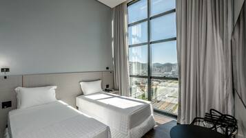 A modern hotel room with two beds, a large window with a city view, a desk, and a chair.