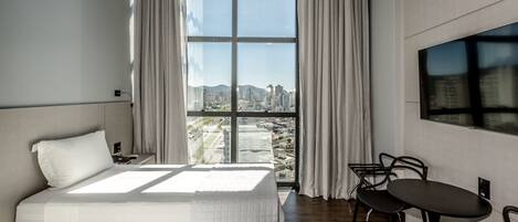 A modern hotel room with a large window, a bed, a desk, and a chair.