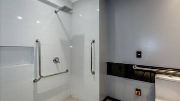 Un baño moderno con azulejos blancos, ducha con cabezal de ducha manual y inodoro con barra de apoyo.