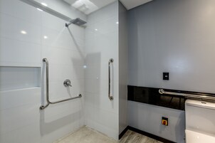 A modern bathroom with white tiles, a shower with a handheld showerhead, and a toilet with a grab bar.