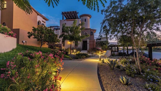 A well-lit pathway leading to a building with a tower, surrounded by landscaped gardens and a pergola.