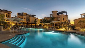 A pool area with lounge chairs and umbrellas, surrounded by buildings with Mediterranean-style architecture.
