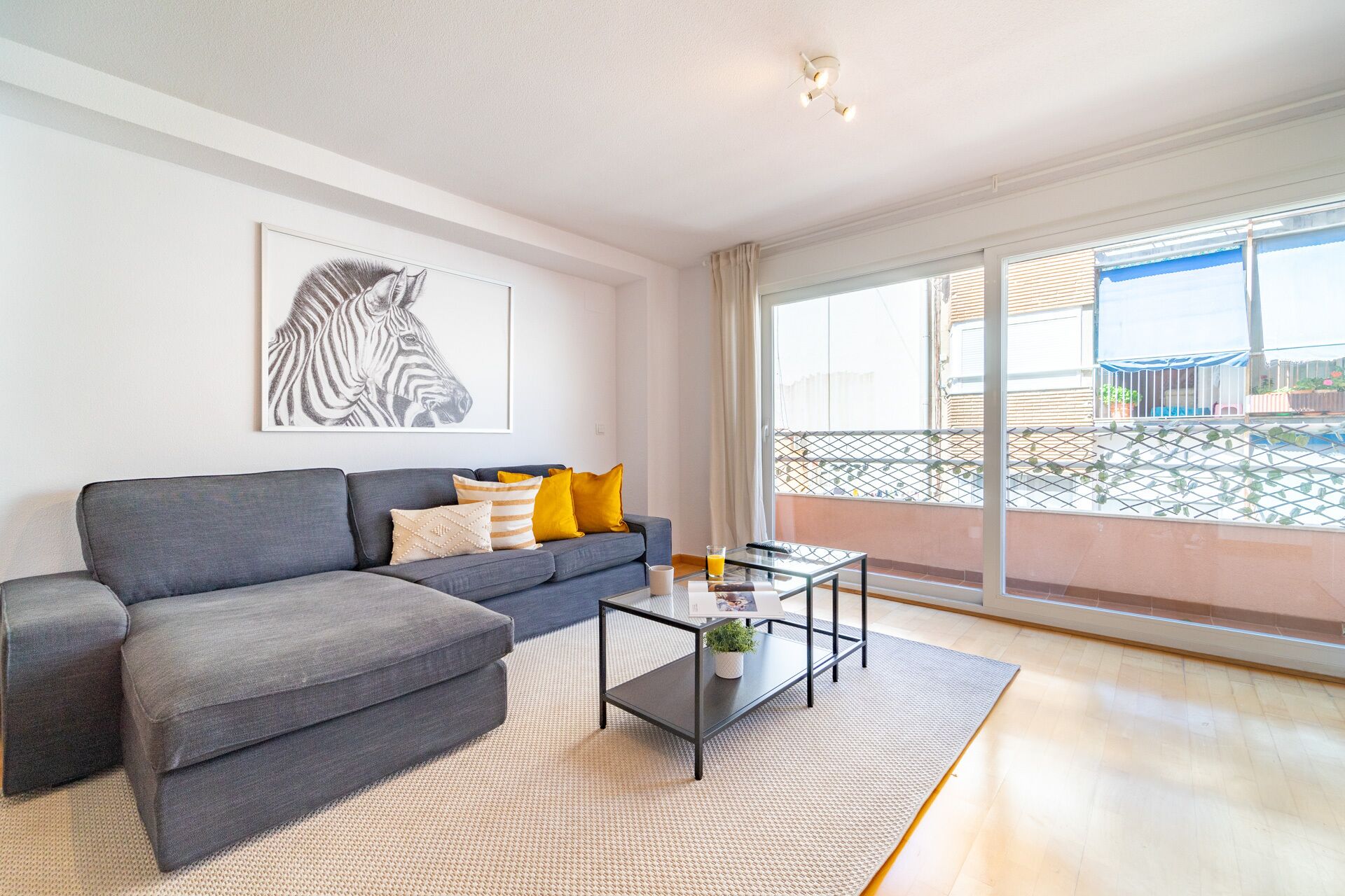 Een woonkamer met een grijze bank, een glazen salontafel en een afbeelding van een zebra aan de muur.