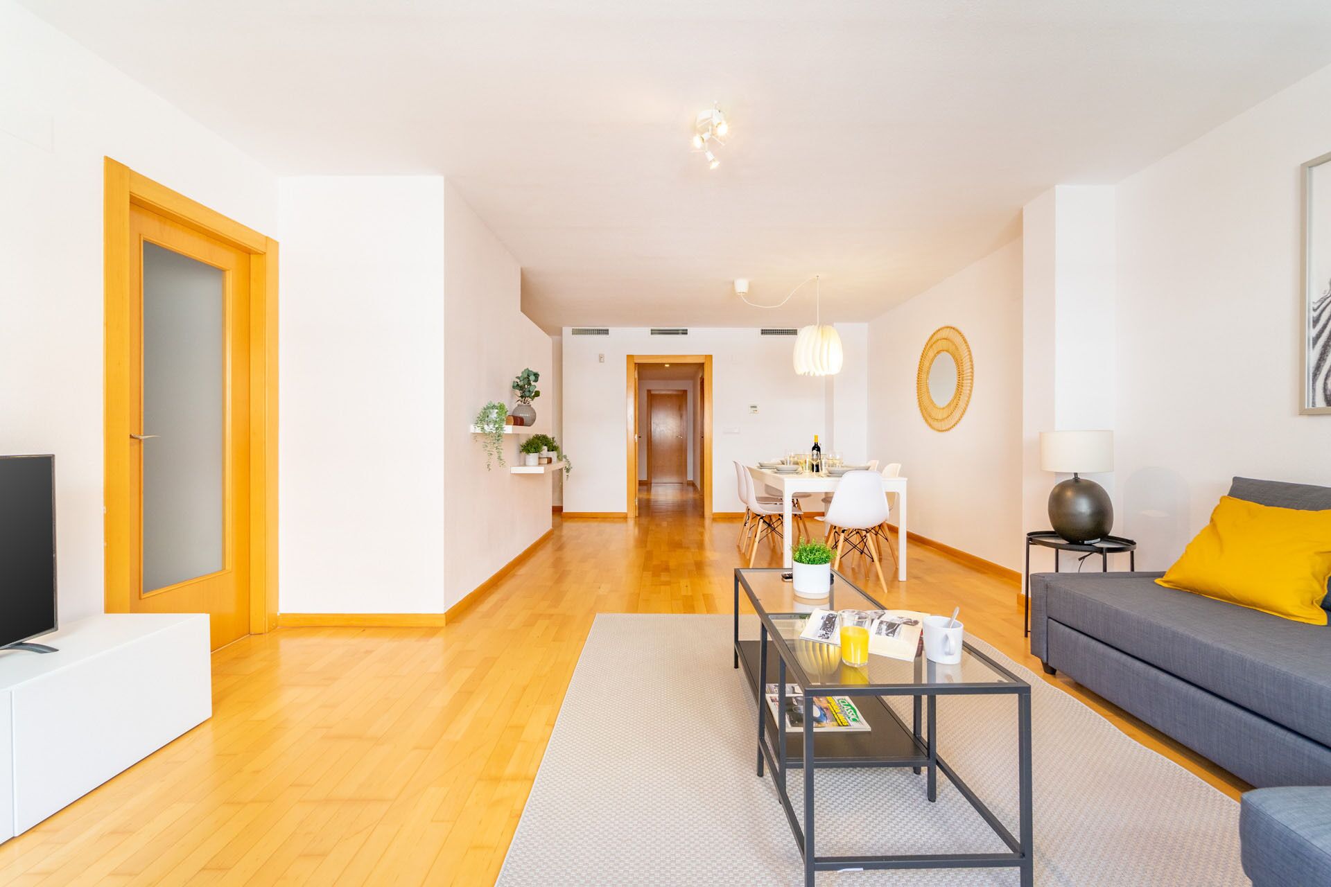 A modern living room with a sofa, coffee table, and TV. There is a dining area with a table and chairs.