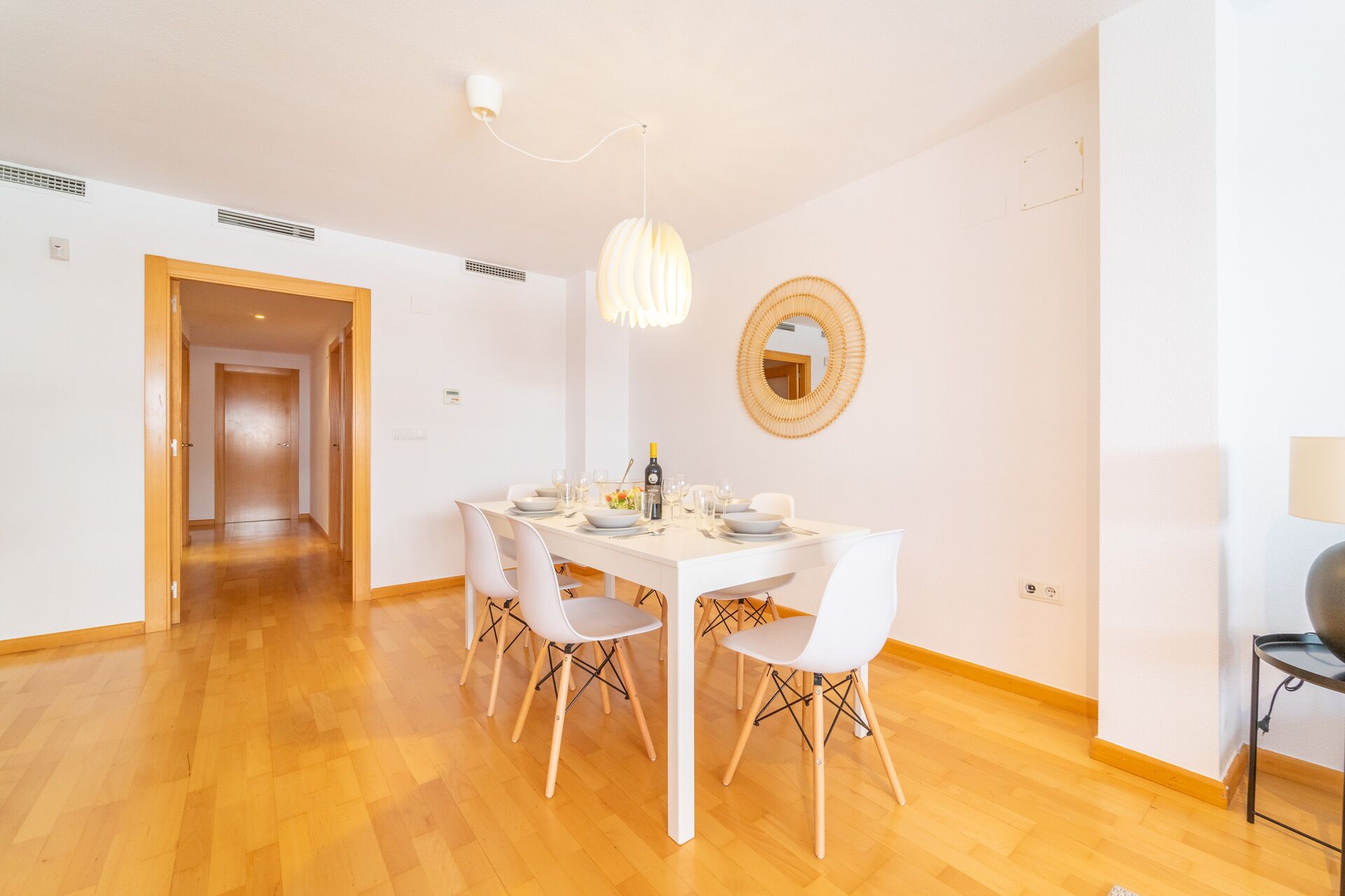 Et spiseområde med et hvidt bord dækket til fire personer, omgivet af hvide stole med træben. En moderne pendellampe hænger over bordet, og et rundt spejl er monteret på væggen.