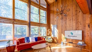 A spacious room with large windows, a red sofa, a wooden coffee table, and a projector screen displaying a logo. - DESC09 - Retro Home, Sunken Living Room, Private Hot Tub (Sunriver)