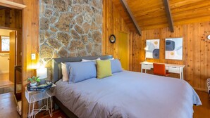 A bedroom with a stone wall, wooden ceiling, a bed with blue and green pillows, a desk with a chair, and abstract wall art. - DESC09 - Retro Home, Sunken Living Room, Private Hot Tub (Sunriver)