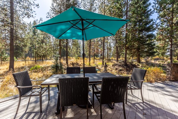 An outdoor dining area with a table and chairs, a large umbrella, and a forested backdrop. - DESC09 - Retro Home, Sunken Living Room, Private Hot Tub (Sunriver)