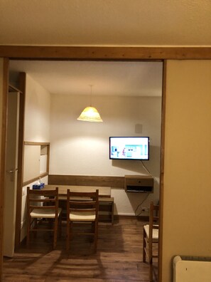 Une pièce comprenant une table à manger en bois, des chaises, un téléviseur fixé au mur et une lampe suspendue.