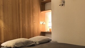 Ein kleiner, gemütlicher Raum mit einer Wand aus Holzlatten, einem Bett mit grauer Decke, einem kleinen Holztisch und einem Regal mit Dekoration.
