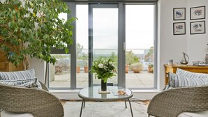 A modern living room with wicker furniture, a glass table with a vase of flowers, and a large sliding glass door leading to a balcony with potted plants. - Putney Rooftops - Two Bedroom Apartment, Sleeps 4 (London)