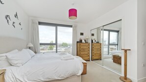 A bedroom with a large bed, a dresser, a mirror, and a view of the city through large windows. - Putney Rooftops - Two Bedroom Apartment, Sleeps 4 (London)
