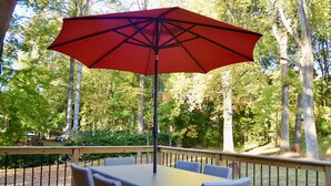 Terraza de madera con mesa y sillas, rodeada por una barandilla de metal, con vista a un área boscosa.
