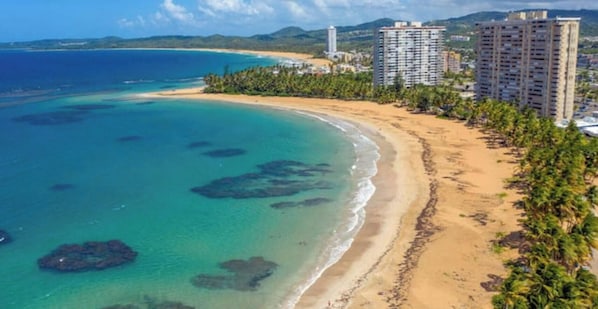Uma área costeira com uma praia arenosa, águas turquesa cristalinas e vários edifícios altos.