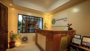 A reception area with a wooden desk, a potted plant, and a sign for Bernardo's Hotel. - Bernardos Lantia Hotel (Basco)