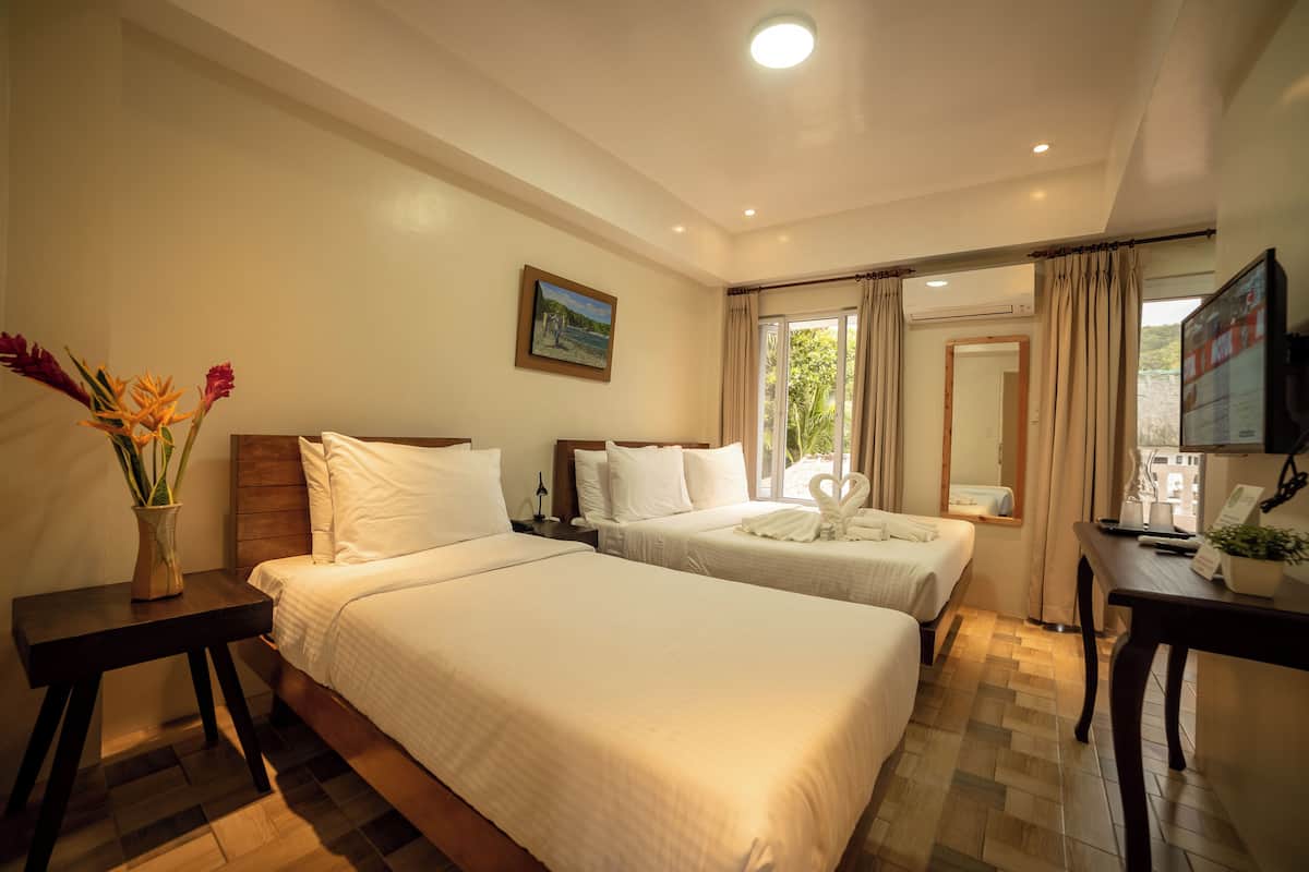 A hotel room with two beds, a wooden headboard, a bedside table with a vase of flowers, a painting on the wall, and a window with curtains.