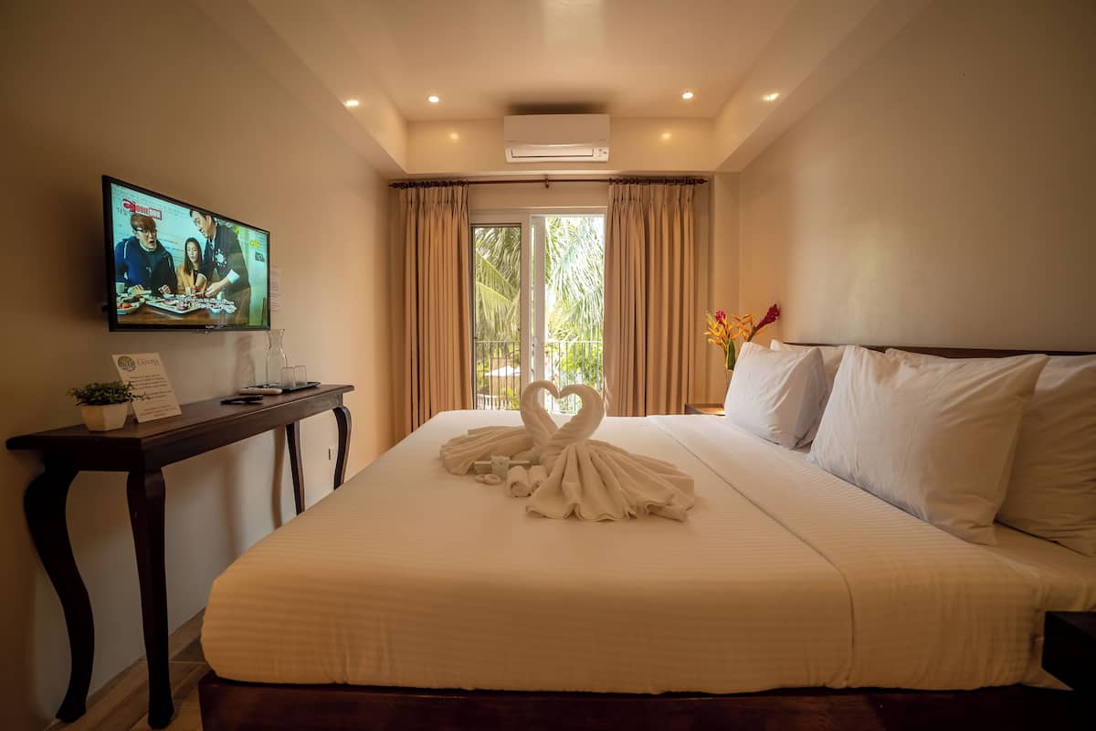 A hotel room with a large bed, a flat-screen TV on the wall, a small table with a plant, and a window with curtains.