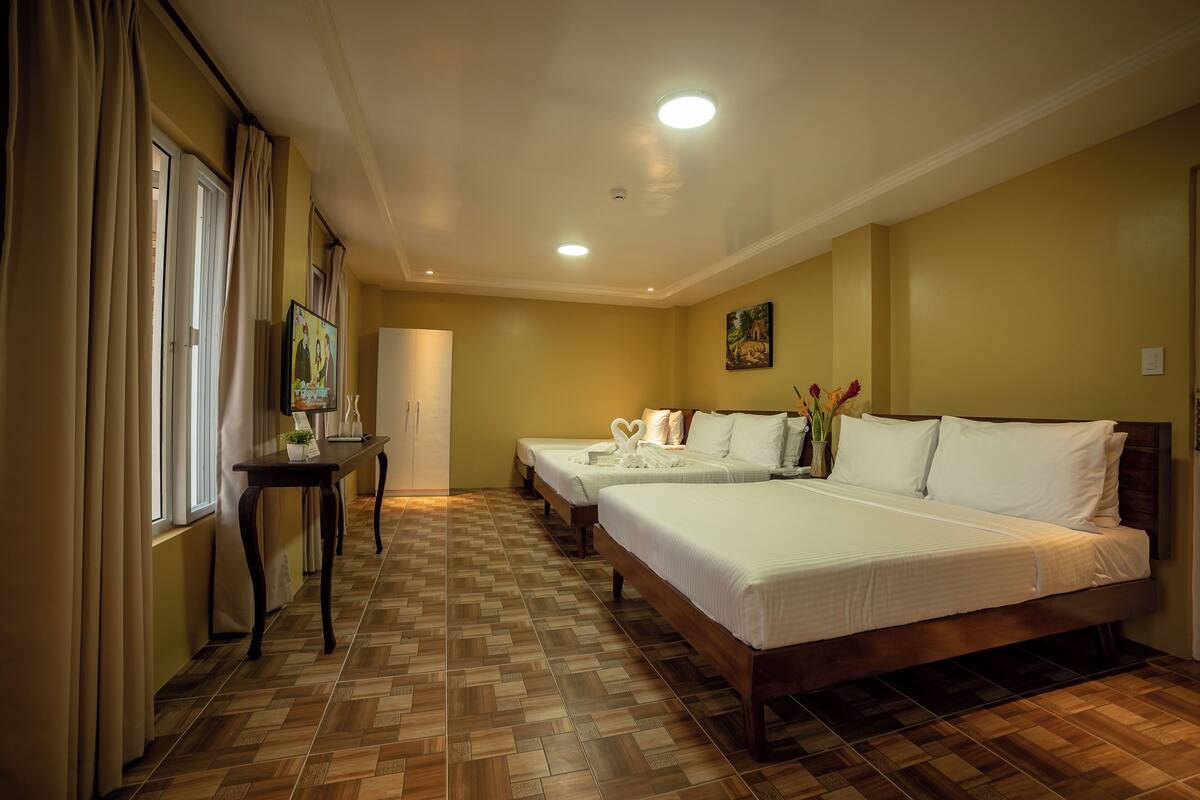 A hotel room with a large bed, two smaller beds, a wooden floor, a desk, and a painting on the wall.