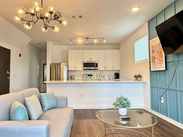 A modern living room with a sofa, a glass coffee table, and a kitchen area with white cabinets and stainless steel appliances.