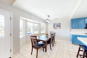 A dining area with a wooden table and chairs, a hanging light fixture, and a window with a view of trees. - Mountain-view home with a game room, washer/dryer, firepit, AC & basketball (Oakhurst)