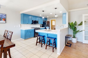 A modern kitchen with blue cabinets, a central island with stools, and a dining area with a wooden table and chairs. - Mountain-view home with a game room, washer/dryer, firepit, AC & basketball (Oakhurst)
