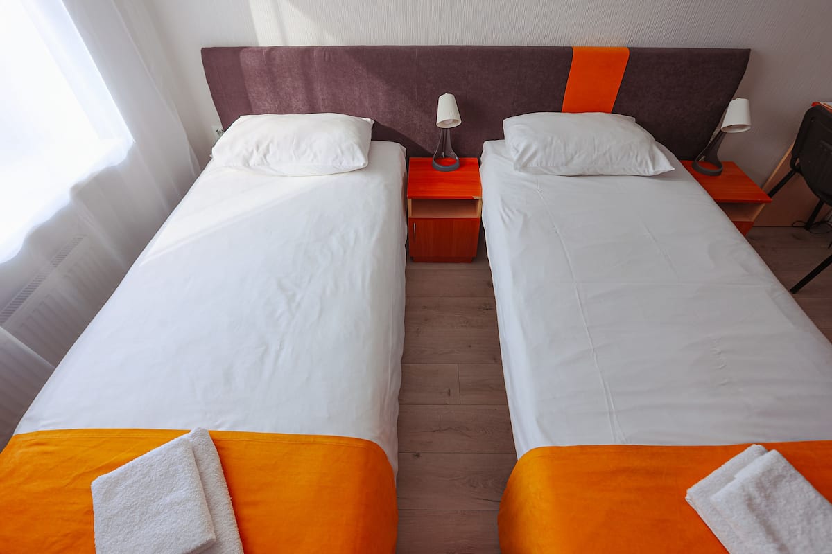 A hotel room with two single beds, each with white linens and orange bedspreads. There are two bedside tables with lamps, and a chair is visible in the corner.