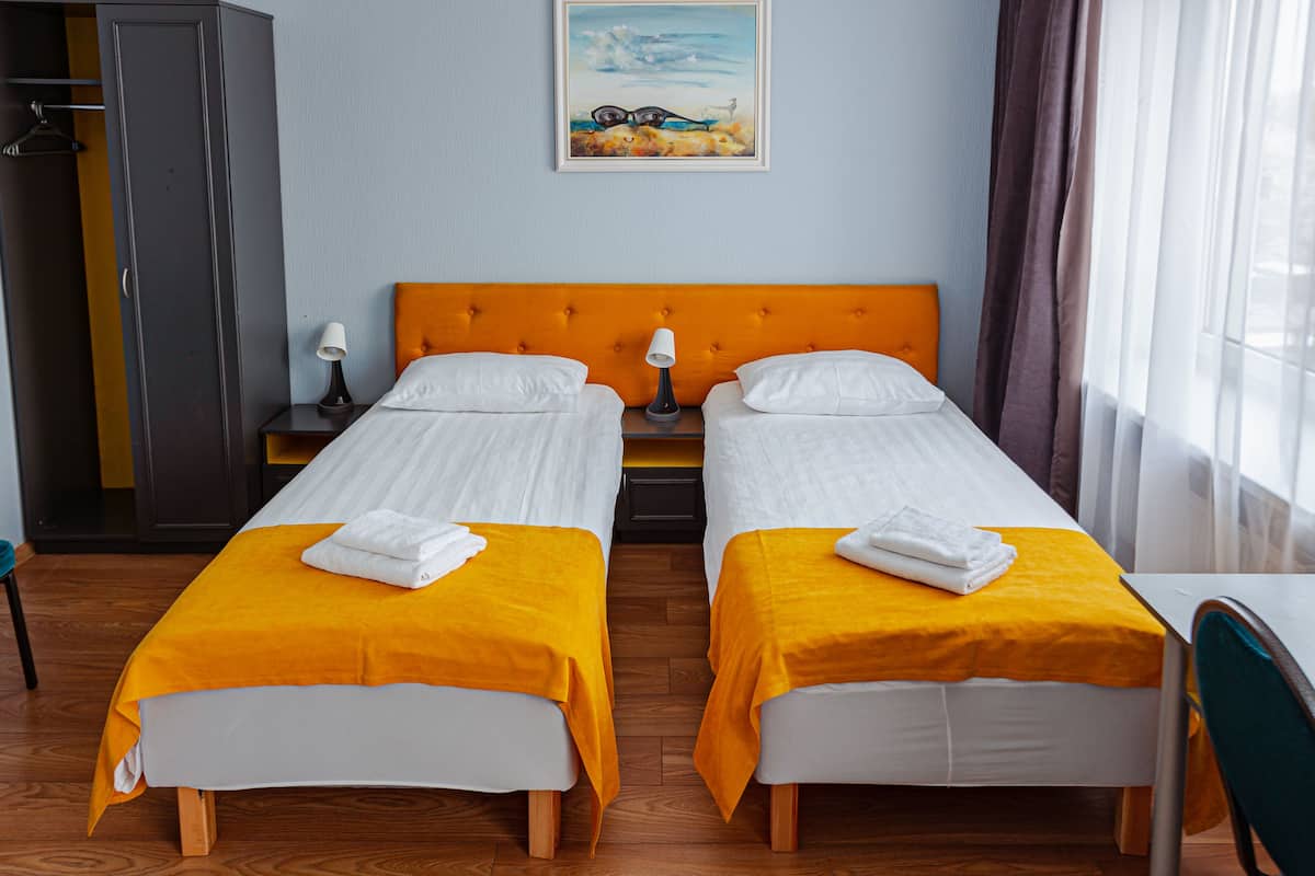A hotel room with two single beds, a wooden floor, a grey wardrobe, a painting on the wall, and a window with curtains.