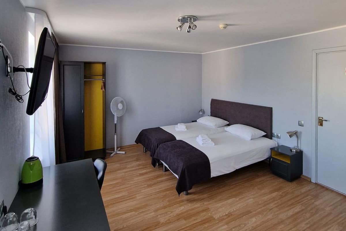 A hotel room with a bed, bedside table, desk, chair, and a small table with glasses and a remote control.