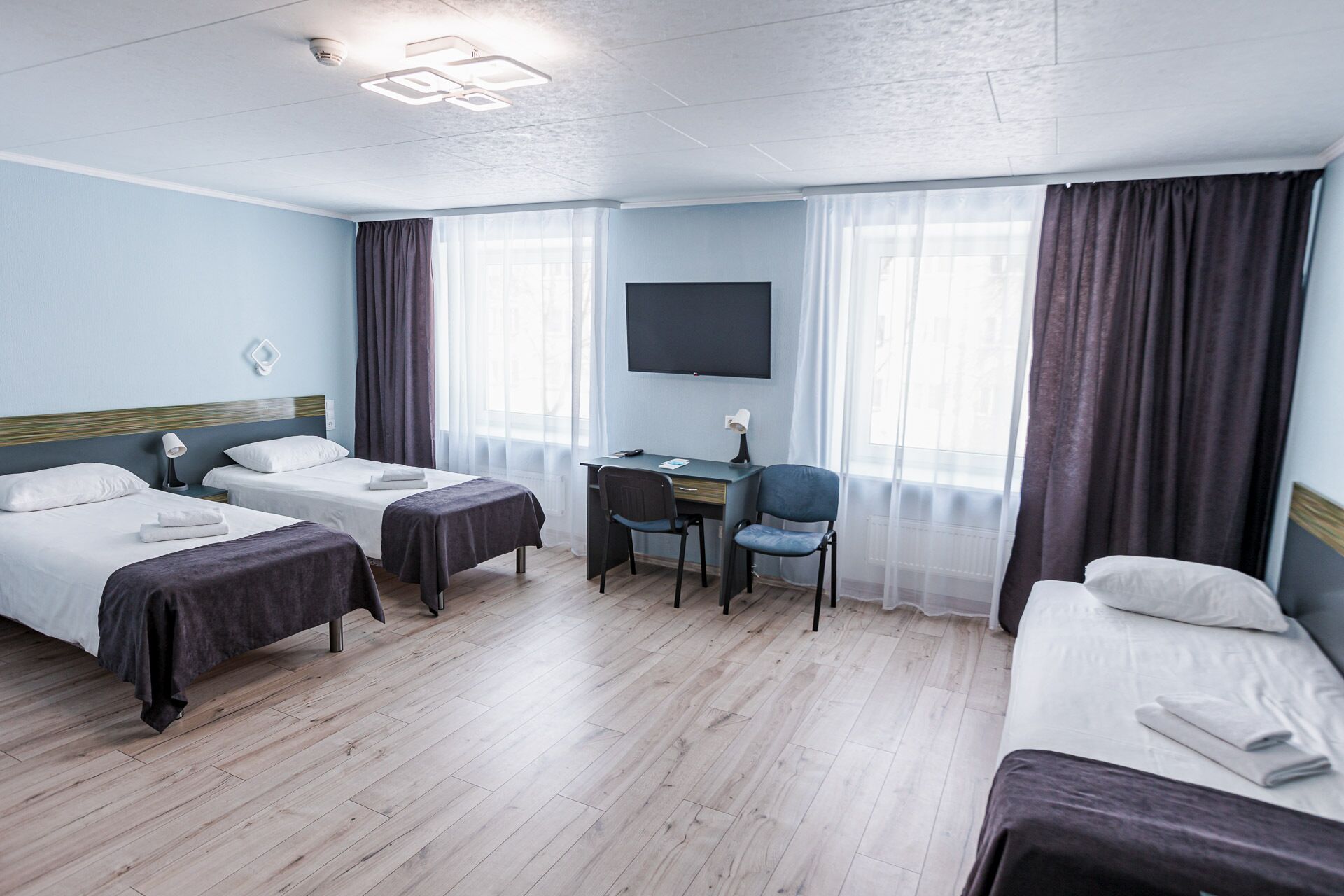 Habitación de hotel con dos camas individuales, un escritorio con silla, un televisor de pantalla plana y un ventanal con cortinas.